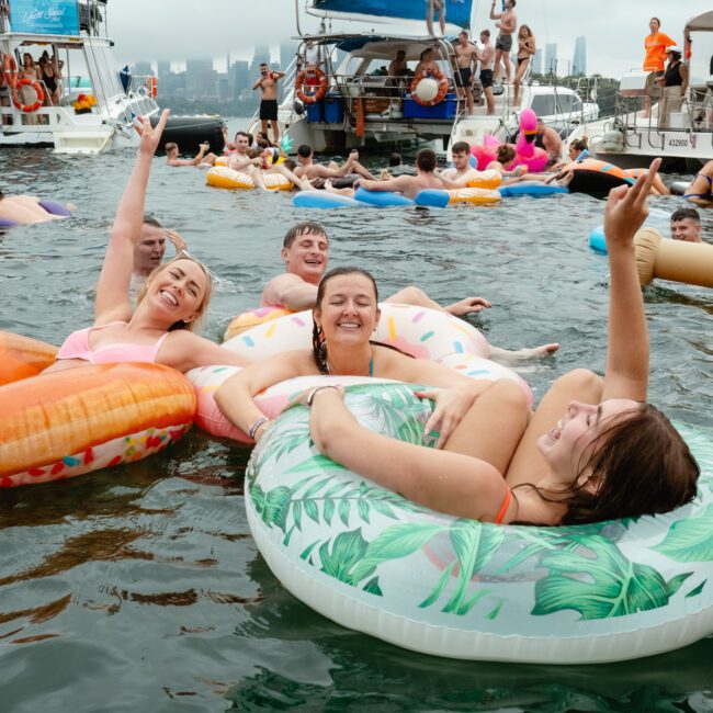 A large group of people float in an ocean bay on inflatable pool toys like donuts, pineapples, and palm leaves. Boats anchored nearby have even more revelers onboard, enhancing the lively, festive atmosphere that everyone seems to be thoroughly enjoying.