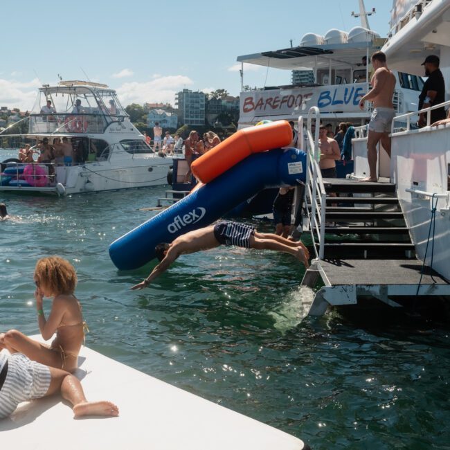 A boat gathering features people swimming and lounging on floaties, while someone uses an inflatable slide from the boat into the water, with a city skyline in the background. Perfect for a luxury yacht hire Sydney or corporate boat events Sydney.