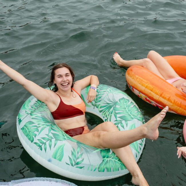 A group of people floating in a lake on colorful inflatable rings. The individual in the foreground is wearing a red bikini with a big smile, raising an arm and stretching a leg. The inflatables feature patterns like leaves, tropical designs, and solid colors.