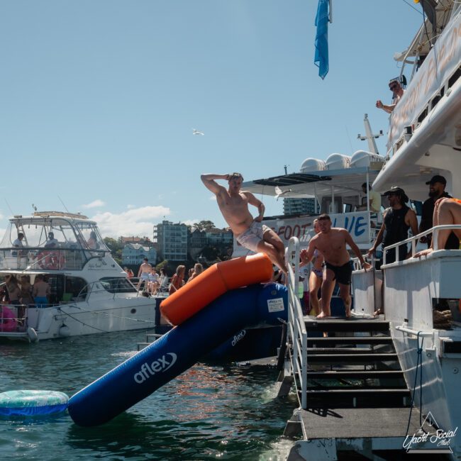 A person in swimwear slides off a blue and orange inflatable slide into the water from a boat, with several other people and boats in the background. It is a sunny day, perfect for a private yacht charter on Sydney Harbour or corporate boat events Sydney.