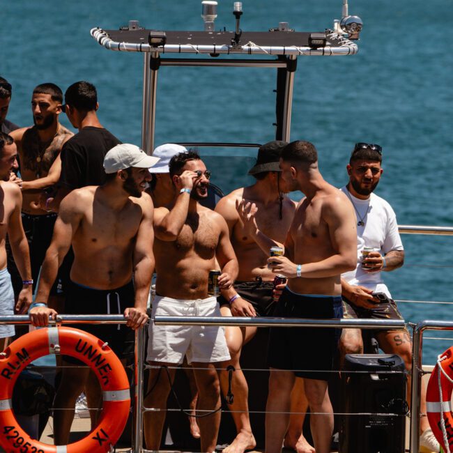 A group of shirtless men is standing and talking on a boat under a sunny sky. The boat cruises on a body of water with scenic views of distant trees and buildings. Several men are holding drinks, while two life preservers and a speaker add to the lively atmosphere on the deck.