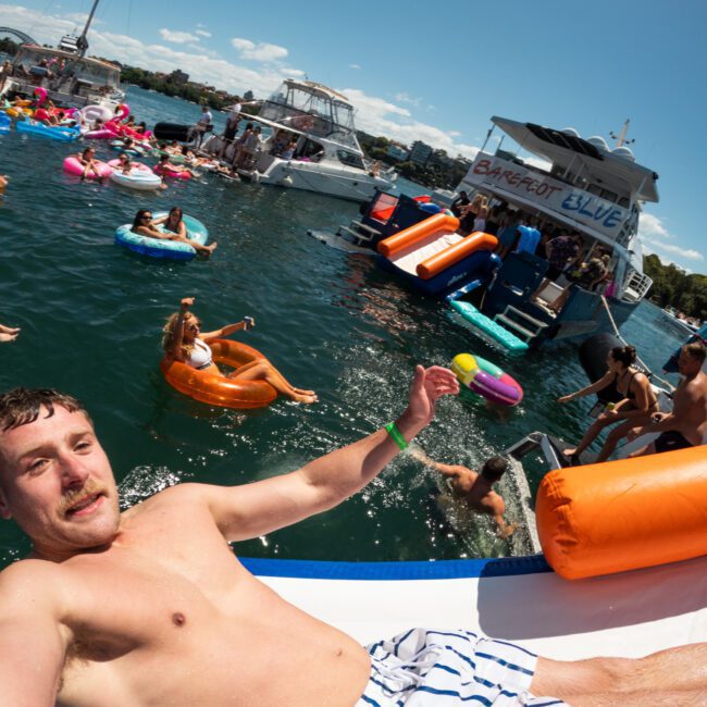A man slides down an inflatable slide into a lake, surrounded by many people on inflatables and boats. Several individuals are lounging on brightly colored floats, enjoying the sunny day. A large boat with a "Bangboat Blue" sign is prominent in the background, adding to the festive atmosphere.