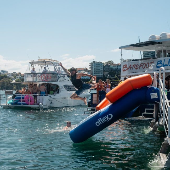 A person jumps from an inflatable slide into the water from a boat, with other people on boats and in the water. A sign reads "BAREFOOT BLUE." Urban buildings and a sunny sky are in the background, creating the perfect setting for a private yacht charter Sydney Harbour.
