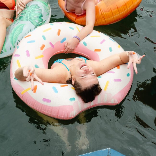 A person beams and makes peace signs with both hands while floating on a sprinkle-decorated donut-shaped pool float in the water. Others relax on various vibrant floats nearby, creating a lively summer scene.