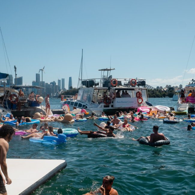 People are floating on inflatable pool toys in the water next to several boats on a sunny day in a coastal city, enjoying the vibrant atmosphere with DJ boat hire Sydney setting the perfect party mood.