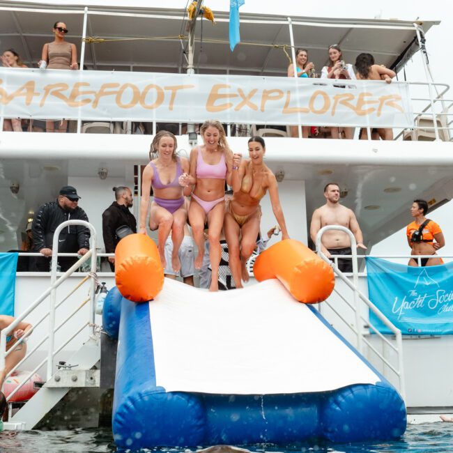 A group of people in swimsuits enjoy a water slide from a boat named "Barefoot Explorer." Three women are at the top of the slide, ready to descend, while others gather around the boat and in the water. A lifeguard is on duty, and banners read "Uptop Adventure Club.