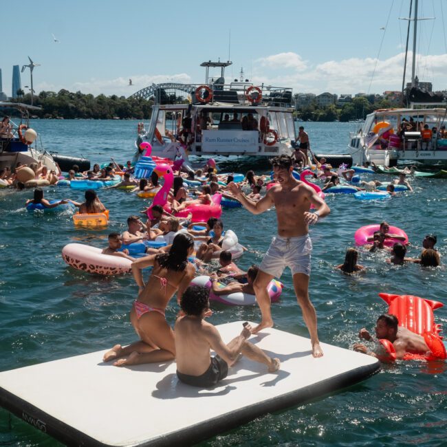 A group of people enjoying a sunny day on various inflatables and an inflatable platform in the water, with boats and onlookers in the background. Perfect for those interested in Sydney boat party hire or luxury yacht hire Sydney.