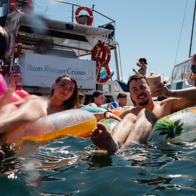 People relaxing on inflatable floats in the water near a boat with a "Rum Runner Cruises" sign on a sunny day. Consider adding some excitement to your outing with DJ boat hire Sydney or opt for luxury yacht hire Sydney.