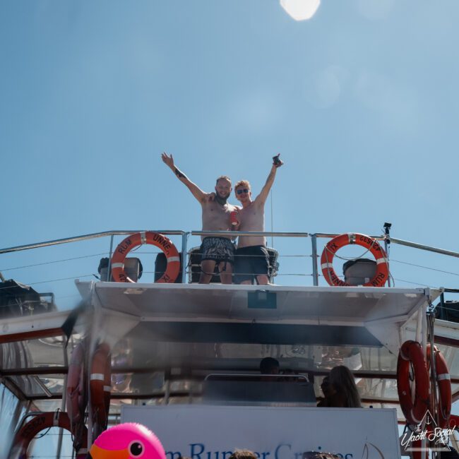 Two shirtless men stand on the upper deck of a boat, raising their arms in celebration during a corporate boat event in Sydney Harbour. Lifebuoys are visible behind them.