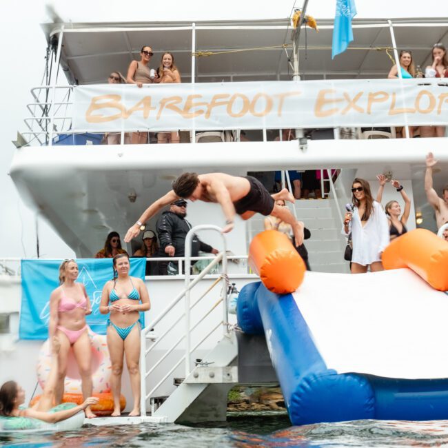 A group of people are having fun on a boat named "Barefoot Explorer." A man slides down a colorful inflatable slide into the water, while others in swimwear stand around or on the boat, watching and laughing. The scene is lively and vibrant.