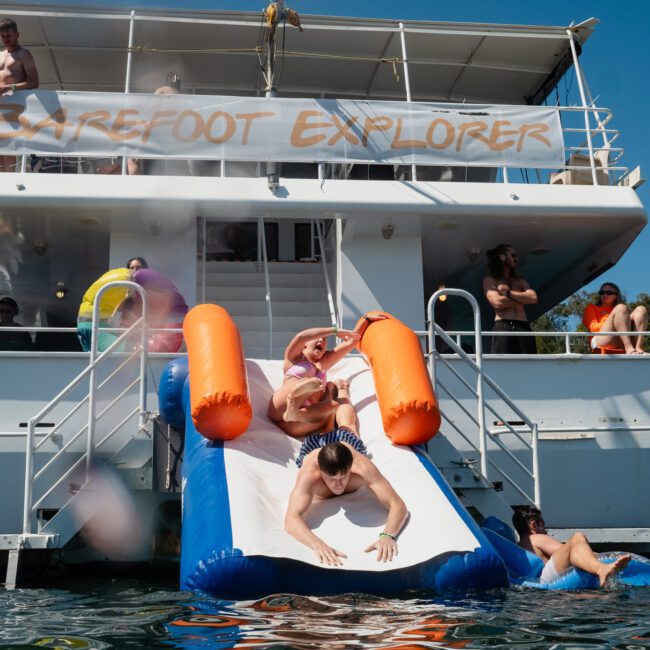 Partygoers slide down an inflatable slide off the back of a boat named "Barefoot Explorer" with others watching from different levels and the water, enjoying a fantastic Sydney boat party hire.