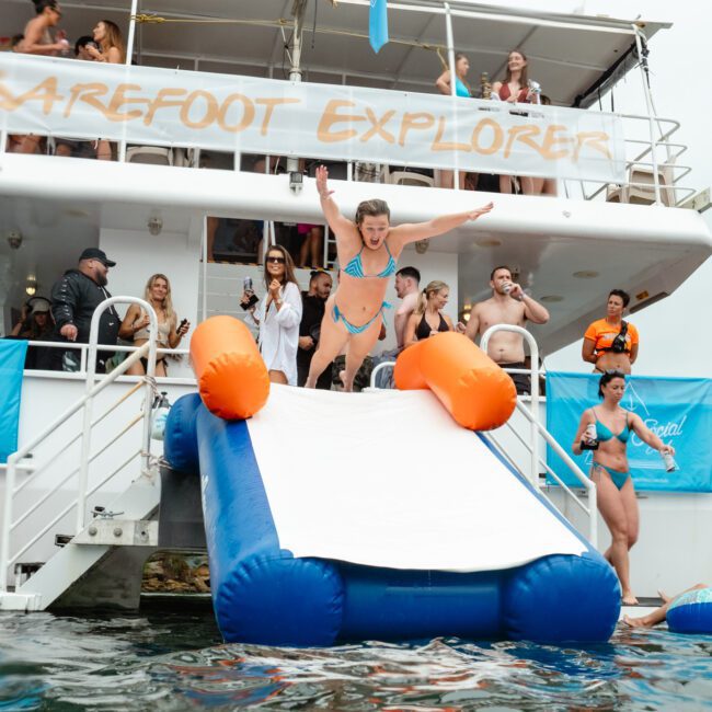 A man in swim trunks is sliding down a large inflatable slide from the deck of a boat into the water, with arms spread wide. The boat, named "Barefoot Explorer," is filled with onlookers and other partygoers in swimsuits enjoying the festive and lively atmosphere.