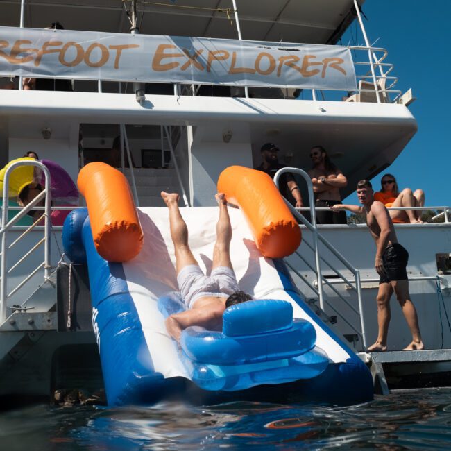 A person slides down an inflatable slide into the water from the back of a yacht labeled "Barefoot Explorer," as several people enjoy the view from the deck during a private yacht charter in Sydney Harbour.