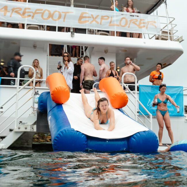 A woman is sliding down an inflatable slide off the back of a boat named "Barefoot Explorer," surrounded by people enjoying the water. Some are on the boat, while others are in the water holding colorful floatation devices. It looks like a joyful and lively scene.