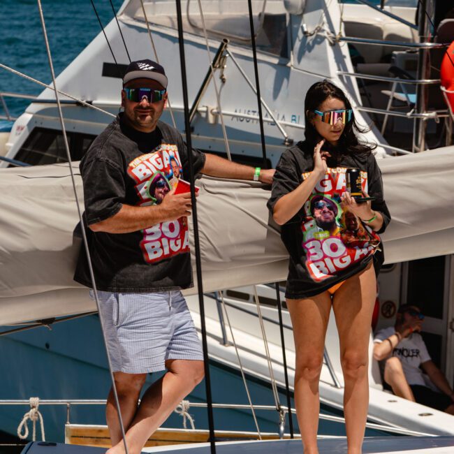 Two people stand on the deck of a boat wearing sunglasses and matching "BIGGIE" T-shirts. One man in a cap and shorts holds a drink, while a woman in shorts enjoys a snack. The boat is docked, offering stunning views of the ocean in the background.