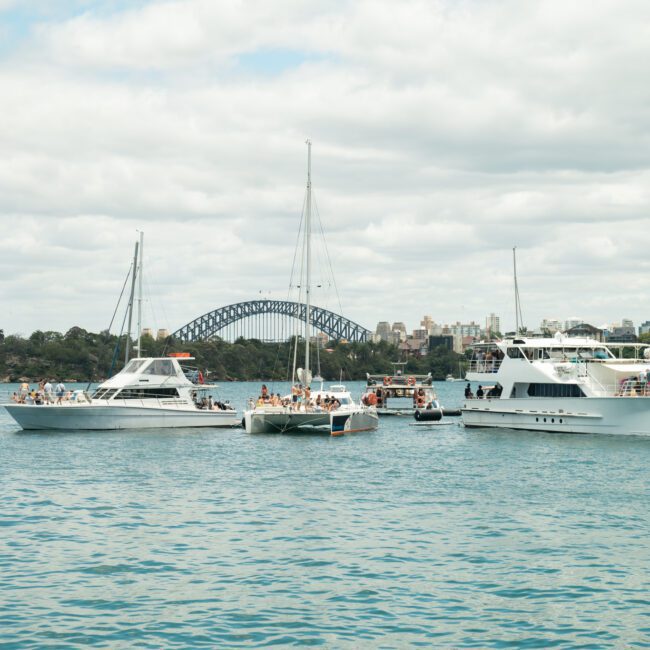 A scenic harbor scene with several boats anchored on calm water. The iconic Sydney Opera House and Sydney Harbour Bridge are visible in the background under a partly cloudy sky, with city buildings lining the horizon, enhanced by vibrant reflections on the water.
