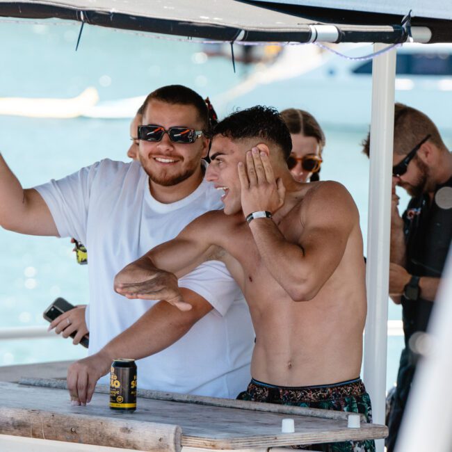 A group of people enjoy a sunny day on a boat. Two men stand at a table, one in a white shirt and sunglasses, smiling and posing enthusiastically, while the other shirtless man gestures energetically. Others in the background laugh and socialize, with the beautiful sea visible behind them.