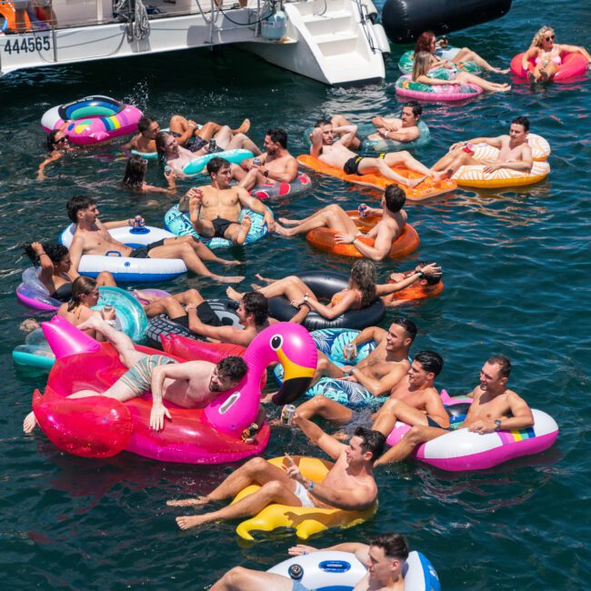 A group of people enjoying a sunny day on the water, floating on colorful inflatable rafts like a flamingo and a unicorn, near a boat. The scene is lively with people socializing and relaxing on the lake.