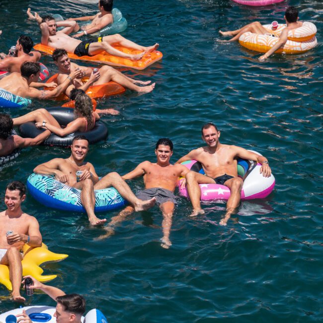 A group of people relax on colorful inflatable pool floats in a vibrant blue body of water. Some floats are shaped like animals, and most people are casually dressed in swimwear. Everyone appears to be enjoying themselves under the sunny sky, creating a picturesque summer scene.