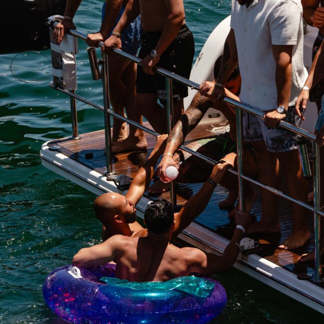 A group of people enjoy a sunny day on a docked boat. Two men are in the water, one in a blue-purple inflatable ring, reaching up to receive a drink from another person on the boat. Others stand on the boat, relaxing and conversing. Logos read "Sail Gustavo" and "Yacht Social Club.