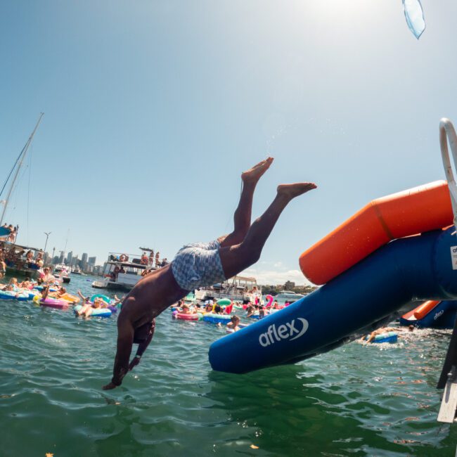 A person in swim trunks dives off an inflatable slide labeled "aflex" and "blue" into clear water amidst other swimmers on floaties. Sailboats and a clear blue sky are in the background, creating a lively and fun waterfront scene reminiscent of The Yacht Social Club events.