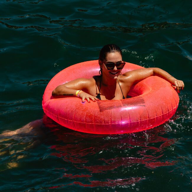 A person with sunglasses smiles while floating in a body of water using a bright pink inflatable ring. The water is dark green, and the sun shines on the person and the ring, creating a cheerful ambiance.