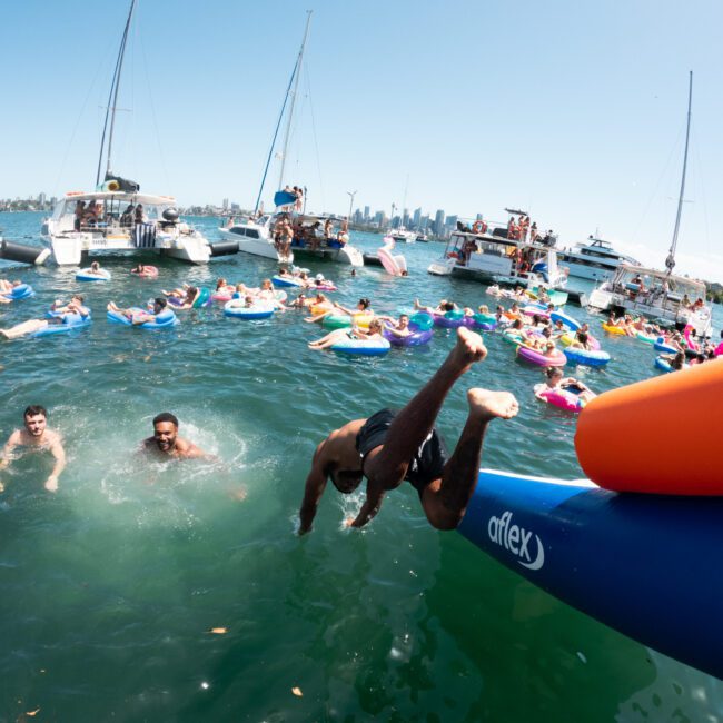 A lively water party features people swimming and floating on inflatables near several anchored boats. One person, mid-dive from an inflatable slide, splashes into the water while others socialize and enjoy the sunny day. The stunning city skyline is visible in the background, enhancing the festive atmosphere.