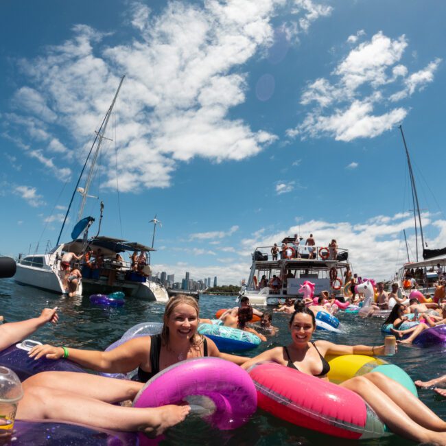 A crowded scene shows people enjoying a sunny day on a lake. Many are floating in colorful inflatable tubes, while boats and yachts are stationed nearby. The backdrop features a bustling city skyline under a clear, blue sky with scattered clouds.