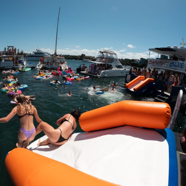 A lively scene of people enjoying a sunny day on the water, featuring multiple boats and colorful inflatable tubes. Two women in bikinis slide down an inflatable slide on a boat, while a crowd watches and participates in the fun. The impressive city skyline is visible in the background.