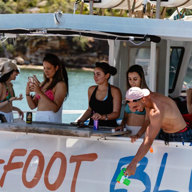 A group of people on a boat named "Barefoot Blue" are enjoying a sunny day. Some are taking photos, while one person is spraying a bottle. The boat is docked near a picturesque area with trees and water in the background, perfect for capturing moments with friends.