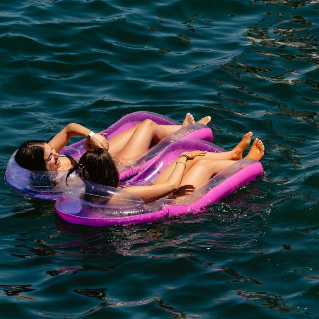 Two people are floating on purple inflatable loungers in a body of water. They are relaxing next to each other, facing slightly towards each other. The water around them is deep blue and calm, with the sunlight reflecting off its surface. The inflatable loungers have transparent headrests for added comfort.