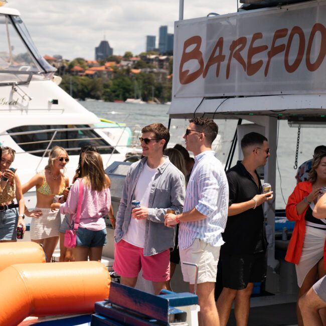 A group of people socializing on a boat named "Barefoot," with a cityscape in the background. They are dressed in casual summer clothing, holding drinks, and appear to be enjoying a sunny day near the water. Bright orange flotation devices add a pop of color in the foreground.