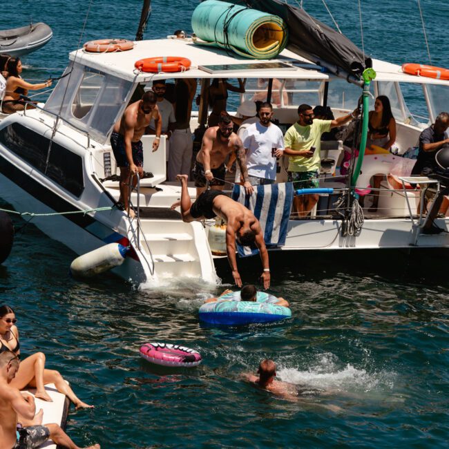 A lively group of people enjoys a sunny day on a boat surrounded by clear blue water. Some are relaxing on the deck, while others swim nearby or float on inflatable rafts. The boat has colorful awnings and safety gear, adding to the fun and festive vibe with a sense of adventure in the air.