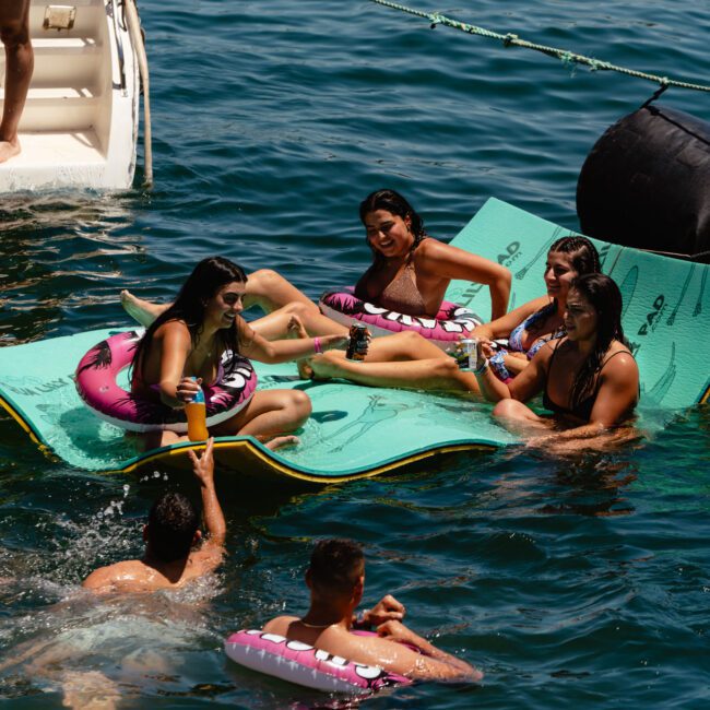 People on a floating mat in the water are smiling and having fun. Some are sitting, and others are in the water holding onto the mat. Two large inflatables add to the scene's vibrancy. The background includes a boat and clear blue water with gentle waves enhancing the picturesque setting.