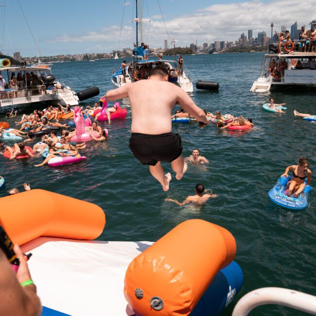 A man jumps off an inflatable water slide into a crowded bay where people are floating on inflatable pool toys. Several boats are docked nearby, accompanied by the soothing sounds of the waves. The city skyline is visible in the background under a partly cloudy sky, creating a festive and lively atmosphere.