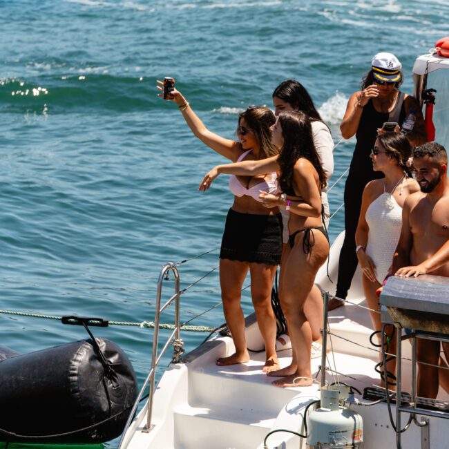A group of people are standing on a boat in the water. Two women at the edge of the boat are taking a selfie while others nearby watch. One person is extending an arm out toward the water, and several people in swimsuits and casual attire are enjoying the sunny moment.