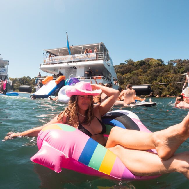 A person lounging on a vibrant inflatable unicorn float in the water, wearing a pink cowboy hat, with boats and others swimming nearby. The scene is bright and sunny, capturing a fun, outdoor gathering or lake party with friends.