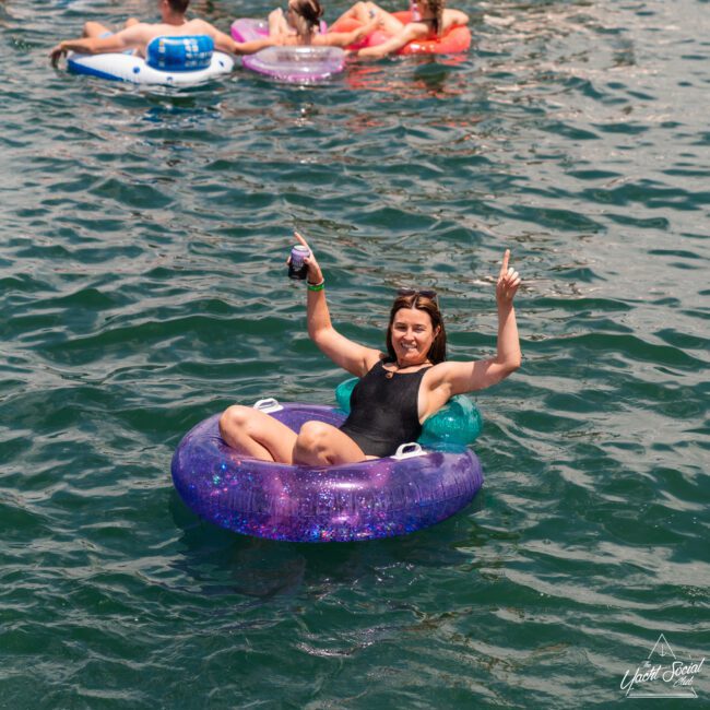 A woman in a black swimsuit floats on a purple inflatable ring in a lake, smiling and raising her hands, one holding a cocktail. Other people in colorful inflatable rings and tubes are visible in the background. The atmosphere is relaxed and joyful.