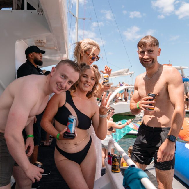 A group of people enjoying a lively boat party. The main focus is on four friends near the boat's edge, smiling and holding drinks. In the background, more people are swimming and relaxing on colorful inflatables in the water under a sunny sky.
