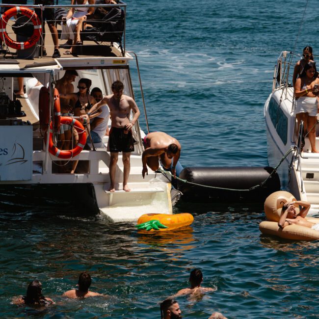 A group of people enjoying a sunny day on boats and in the water. Some are lounging on the boat, others are swimming, and a few are floating on colorful inflatables. The boats are anchored close to each other in a picturesque ocean setting with laughter and splashes filling the air.