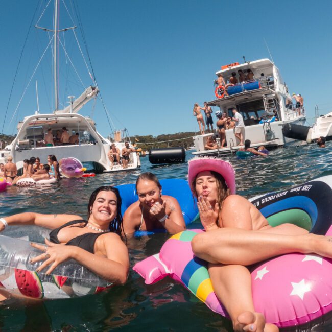 A group of people enjoying a sunny day on the water. Three individuals are floating on inflatable rings, with boats and more people in the background. The atmosphere is lively and vibrant, featuring a fun social gathering with various watercraft seamlessly blending into the scene.