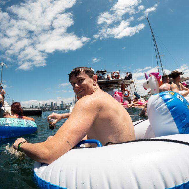A group of people enjoying a sunny day on the water surrounded by various inflatable pool floats, including a whimsical unicorn float. The person in the foreground is smiling and sitting in a white and blue inflatable ring, holding a refreshing drink. A stunning city skyline is visible in the background.