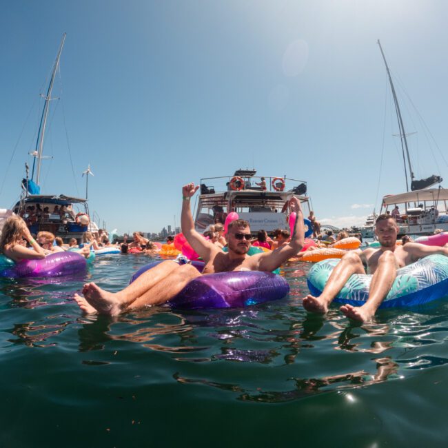 People enjoying a sunny day on the water, floating on colorful inflatable rafts near boats. Some are waving and raising their arms with clear skies and a bright atmosphere. Sailboats glide gracefully in the background, creating an idyllic scene.