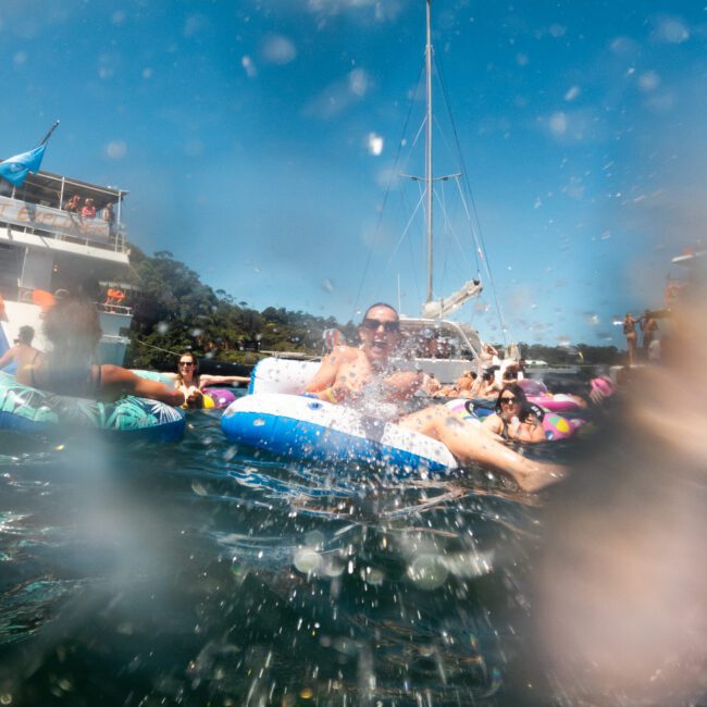 People are joyfully lounging on inflatable tubes and swimming in the water beside a boat. The scene is vibrant with sunny skies and multiple boats in the background. The playful water splashes add a dynamic element to the fun, lively atmosphere, with paddleboards gliding gracefully nearby.