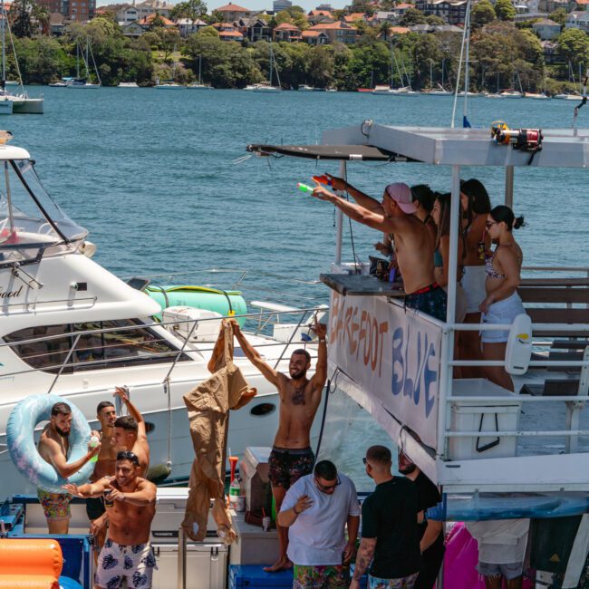 A lively scene on a sunny day featuring a group of people enjoying a boat party. Some are on the boat in colorful swimwear, engaging in water gun fights and vibrant conversations. The backdrop includes a serene waterfront view with city buildings and lush trees visible.