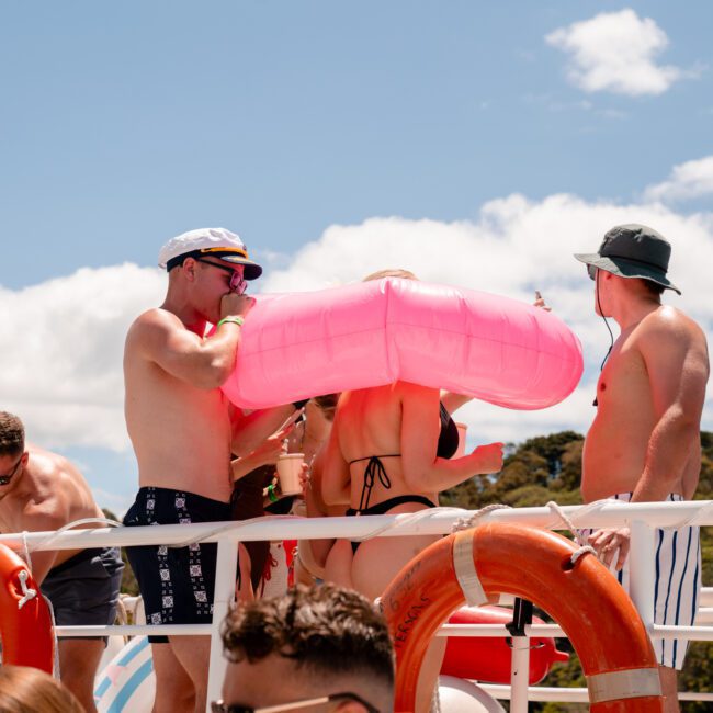 A group of people enjoying a sunny day on a boat, with one person wearing an inflated pink float around their torso. They are in swimwear, surrounded by life rings and a railing. The sky is blue with some clouds, and the scenic landscape includes rolling hills in the background.
