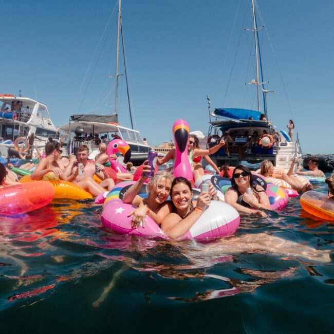 A lively group of people enjoying a sunny day on the water, floating near boats. Many are on inflatable pool floats of various shapes and colors, including a pink flamingo and an eye-catching unicorn float. The atmosphere is festive with smiles and raised drinks.