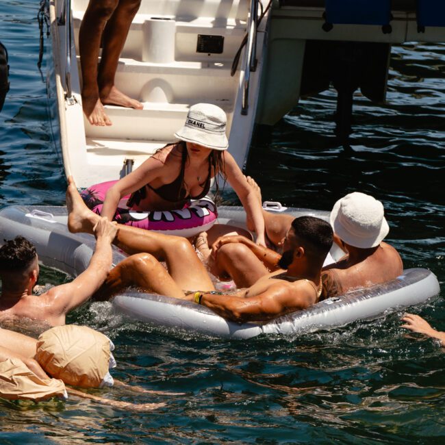 A group of people enjoying a sunny day on the water. Several individuals are floating on colorful inflatable rafts and swimming near the rear of a boat. One person on the boat is helping friends aboard. The setting is relaxed and fun, making for an unforgettable day under clear skies.