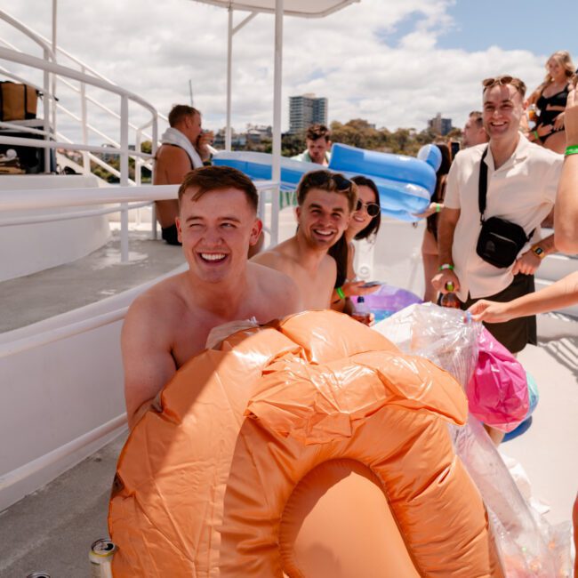 A group of young adults enjoy a sunny day on a boat, smiling and preparing colorful inflatable floats. One person in the forefront holds an orange float, while others in swimsuits and casual attire interact and laugh joyfully in the background.