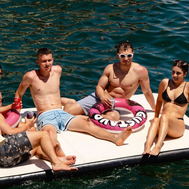 Six people are relaxing on a floating platform on the water. Three are sitting, two are lying down on floaties, and one is handling drinks. All are wearing swimsuits and sunglasses. The dark green water surrounds them, with the "Sail Gustavo" logo on the bottom left corner, adding a touch of elegance.